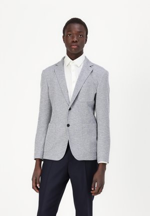Hellgrauer, texturierter Blazer mit Reverskragen, zwei Fronttaschen und schwarzen Knöpfen, getragen über einem weißen Hemd und navyblauen Hosen.