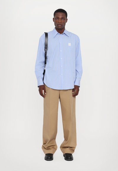 Camisa de botones a rayas azul y blanca, con un corte relajado, combinada con pantalones de pierna ancha en color beige y zapatos negros. Presenta un logo en el pecho.