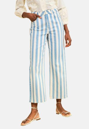 Žena v širokých nohaviciach s modro-bielymi zvislými pruhmi, krémovej vyšívanej blúzke a béžových sandáloch s remienkami na nízkom podpätku.