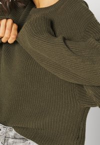 Olivgrüner, gestrickter Pullover mit strukturiertem Muster, weiter Passform und breitem Halsausschnitt. Mit umgeschlagenen Kanten an Ärmeln und Saum.