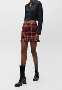 Chemise noire courte à fermeture par boutons, associée à une mini-jupe écossaise rouge à franges au bas. Bottes noires brillantes à talons blocs montantes jusqu'aux genoux.