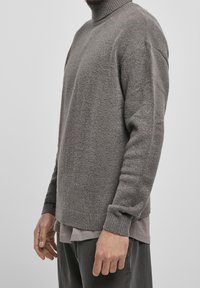 Mann in grauem Rollkragenpullover über einem grauen Hemd und dunklen Hosen, stehend vor einem schlichten Hintergrund.