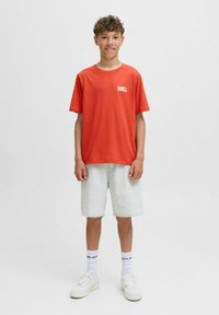 Camiseta de algodón roja con un logo blanco de "SAINT TROPEZ". Combinada con pantalones cortos de mezclilla claros y zapatillas blancas. Fondo liso, vista frontal.