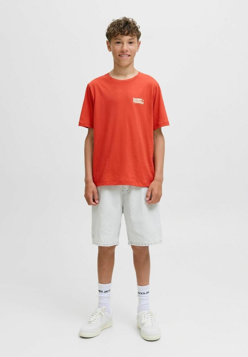 Camiseta de algodón roja con un logo blanco de "SAINT TROPEZ". Combinada con pantalones cortos de mezclilla claros y zapatillas blancas. Fondo liso, vista frontal.