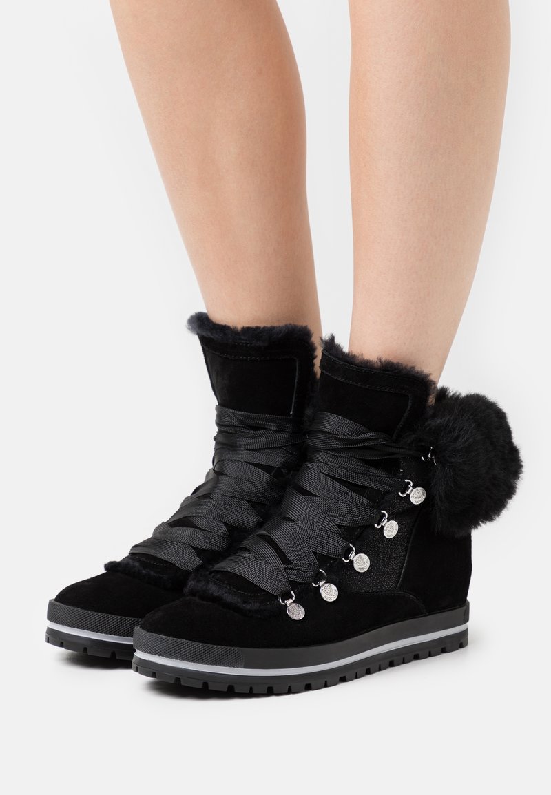 Bottes d'hiver noires doublées de fourrure, avec des lacets épais, des œillets métalliques et un détail pompon, portées sur des jambes nues sur fond blanc.