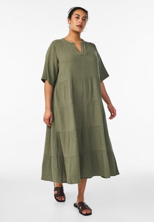 Kvinde iført en ankellang, løs olivengrøn kjole med korte ærmer og sorte åbne slip-on sandaler, stående mod en hvid baggrund.