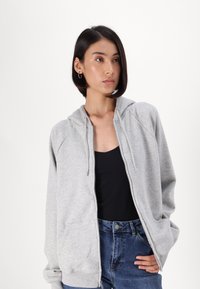 Ανοιχτό γκρι ζακέτα με φερμουάρ και μπροστινή τσέπη, κουκούλα με κορδόνια και ραγλανικά μανίκια, φορεμένη πάνω από μια μαύρη μπλούζα και συνδυασμένη με μπλε τζιν.