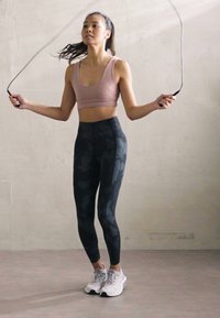 Corda de saltar em uso por uma pessoa vestindo um top curto rosa claro e leggings camuflados pretos, com ténis cinzentos, num chão claro.