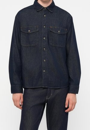 Mand iført mørkeblå denim skjorte med knapper og to forreste brystlommer med knapper samt matchende mørkeblå denim bukser.