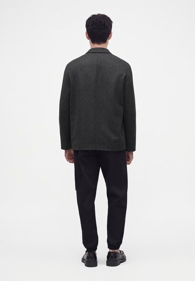 Homme debout de dos, portant une veste à carreaux gris foncé, un pantalon noir et des chaussures noires sur un fond blanc uni.