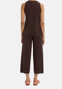 Brązowy bezrękawnik w zestawieniu z pasującymi szerokimi spodniami; gładka faktura; noszone z pomarańczowymi sandałami; minimalistyczny design.