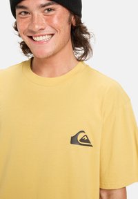 Žluté bavlněné tričko s krátkými rukávy, s černým logem na levé hrudi. Měkký povrch, klasický design s kulatým výstřihem.