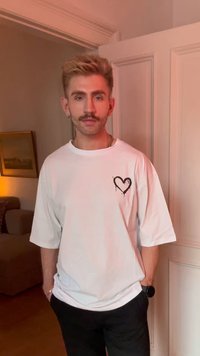 Mężczyzna z wąsami, ubrany w biały, oversize'owy t-shirt z czarnym wzorem serca, stojący w pomieszczeniu przy białych drzwiach, z rękami w kieszeniach.