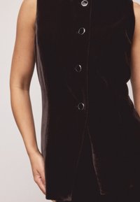 Mustast sametist varrukateta vest, mille ees on kuusnurksete hõbedaste nuppudega ja siidine, elegantne lõige. Sile tekstuur ja keha järgiv disain.