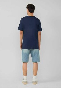 Marineblaue Baumwoll-T-Shirt mit kurzen Ärmeln, kombiniert mit hellblauen Jeansshorts. Weiße Socken und legere beiges Sneakers runden das Outfit ab.