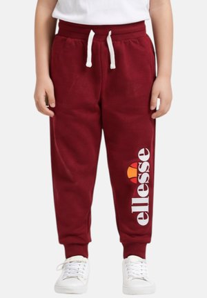 Enfant portant un pantalon de jogging bordeaux avec un cordon blanc, des baskets blanches et un logo blanc « ellesse » sur la jambe gauche.