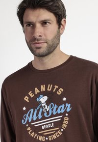 Camiseta de manga larga marrón con un gráfico de Snoopy llevando una gorra azul, con texto crema que dice "PEANUTS All Star Beagle."
