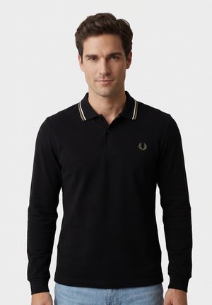 Uomo con capelli castani corti che indossa una polo nera a maniche lunghe con colletto rigato bianco e jeans azzurri, in piedi davanti a uno sfondo chiaro e uniforme.