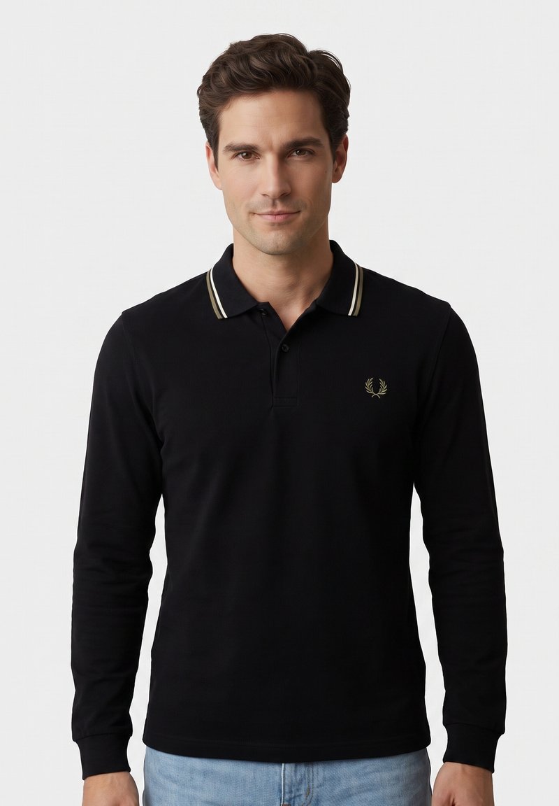 Homme aux cheveux courts et bruns portant un polo noir à manches longues avec un col rayé blanc et un jean bleu clair, debout devant un fond uni et clair.