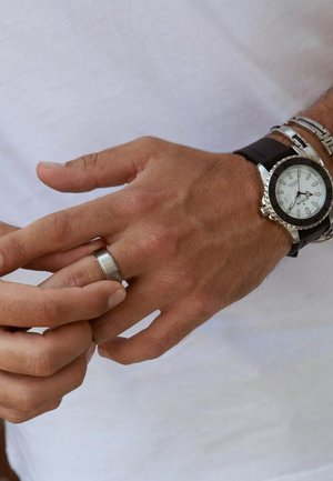 Silver wristwatch with a white face and black bezel, paired with a silver ring. Hand with light skin tone and short nails, wearing a loose white shirt.