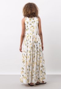 En kvinde med krøllet hår har en ærmeløs, lagdelt maxikjole med et blomsterprint i gul og grøn, mens hun står op ad en ensfarvet baggrund.