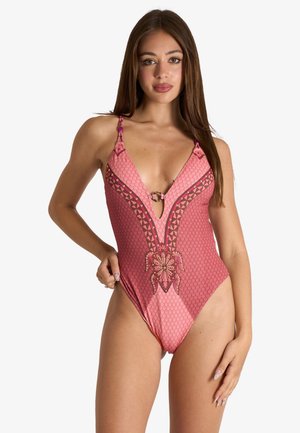 Giovane donna con lunghi capelli castani che indossa un costume intero rosa con motivo a fantasia e scollatura a V profonda, in piedi davanti a uno sfondo bianco.