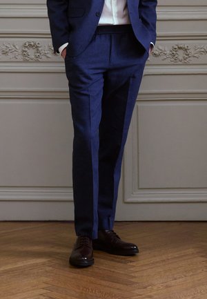 Pantaloni tailored blu navy con una texture liscia, davanti piatto e tasche laterali abbinati a scarpe eleganti in pelle marrone su un pavimento in legno.