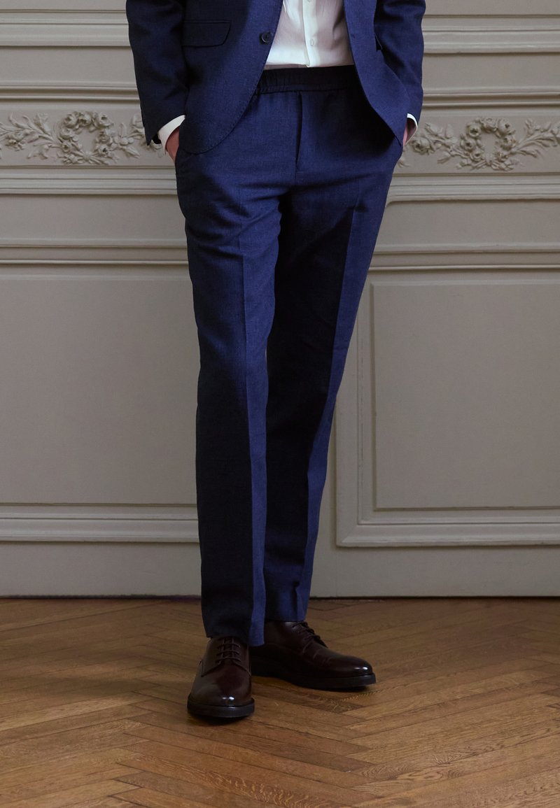 Mornarsko krojene hlače z gladko teksturo, ravnim sprednjim delom in stranskimi žepi, v kombinaciji s celo usnjenimi čevlji rjave barve na lesenem tleh.
