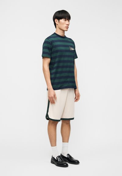 Ριγέ βαμβακερό μπλουζάκι σε πράσινο και ναυτικό χρώμα με τσέπη, σε συνδυασμό με μπεζ σορτς με πράσινες λεπτομέρειες. Το μοντέλο φοράει μαύρα μοκασίνια και λευκές κάλτσες.