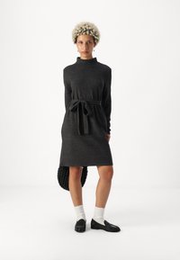 Γκρι πλεκτό φόρεμα με ψηλό λαιμό και μακριά μανίκια, διαθέτοντας ζώνη που δένει στη μέση. Συνδυάζεται με μαύρες loafers και λευκές κάλτσες.