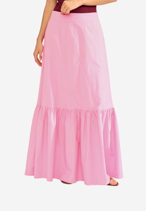 Femme portant une longue jupe rose clair avec un ourlet à volants froncés, associée à un haut rouge foncé et des sandales marron.