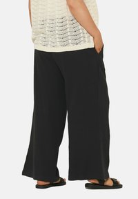 Zwarte wijde broek van textuurstof, met zijzakken en een relaxte pasvorm, gecombineerd met zwarte sandalen.