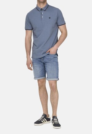 Blaues Poloshirt mit kontrastierendem Kragen, kombiniert mit aufgerollten Denimshorts. Die Schuhe sind schwarz mit weißen Streifen. Sauberer, lässiger Look.
