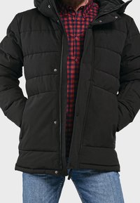 Giacca imbottita nera con zip e chiusura a scatto, dotata di cappuccio e due tasche laterali, indossata sopra una camicia a quadri rossi e neri.