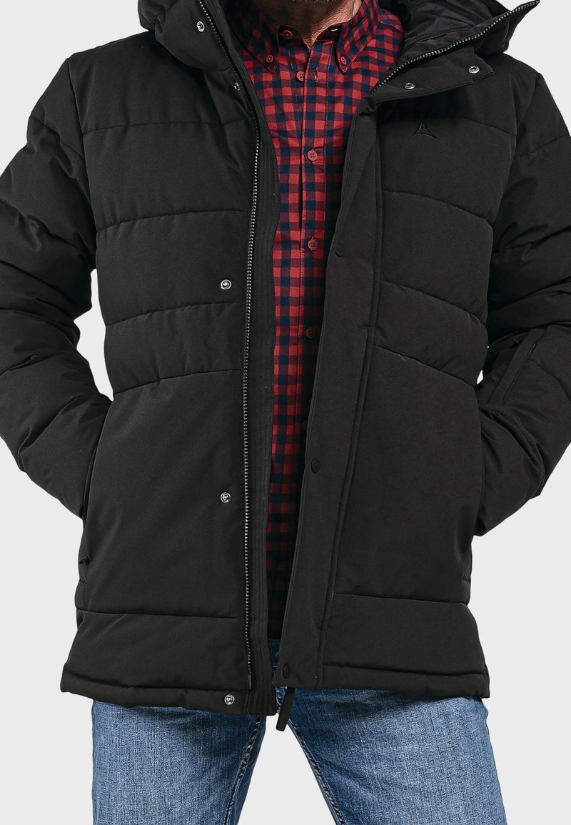 Giacca imbottita nera con zip e chiusura a scatto, dotata di cappuccio e due tasche laterali, indossata sopra una camicia a quadri rossi e neri.