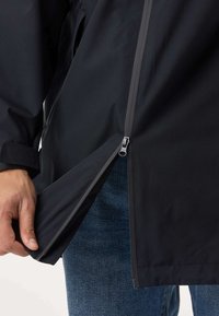 Ruka drží otvorený spodný zips čiernej bundy oblečenej na modrých džínsoch.