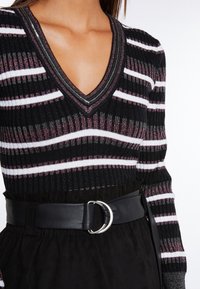 Haut en maille rayé noir et blanc avec des accents métalliques, présentant un col en V et une texture côtelée. Associé à une large ceinture en cuir noir avec une boucle argentée en forme d'anneau.