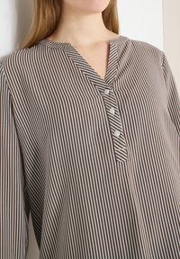 Femme portant un chemisier à rayures verticales marron et blanches avec un col fendu et trois boutons blancs sur le devant.