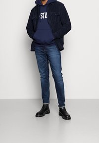 Chaqueta de pana azul marino sobre una sudadera con texto azul marino. Pantalones vaqueros de corte ajustado y botas de cuero negras hasta el tobillo completan el conjunto.