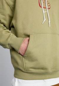 Tamsiai žalia džemperis su priekinėmis kišenėmis, baltos raišteliai ir raudona grafika ant krūtinės. Minkšta medvilnės mišinio tekstūra.