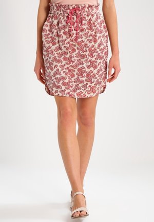 Kobieta ubrana w spódnicę do kolan w czerwony wzór paisley i różowy sznurek, w parze z beżowymi sandałami z odkrytymi palcami na białym tle.