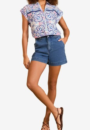 Kobieta w bezrękawnym koszuli z kołnierzykiem z niebiesko-różowymi wzorami, wysokim stanem szortów jeansowych i brązowych sandałach z paskami, pozująca z ręką w kieszeni.