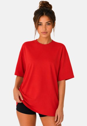 Jonge vrouw met haar in een knot draagt een oversized rood T-shirt en zwarte korte broek, staand tegen een effen lichte achtergrond.