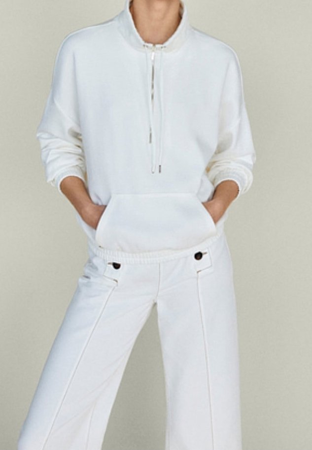 Persona che indossa un pullover bianco a mezza zip con tasca frontale e pantaloni bianchi a vita alta con tasche laterali abbottonate. Mani nelle tasche.