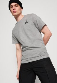 Tricou gri din bumbac cu un logo Jumpman negru pe partea stângă a pieptului, purtat cu pantaloni cargo negri. Materialul pare moale și casual.