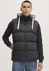 Chaleco acolchado negro con cierre de cremallera, capucha gris con cordones, mangas de punto negro texturizadas y acento de logo de cuero marrón.