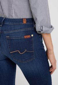 Jeans de mezclilla en azul oscuro con una textura suave. Presenta una etiqueta marrón en el bolsillo trasero y costuras decorativas marrones en un patrón ondulado.