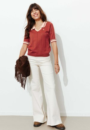 Femme portant un haut en maille manches courtes couleur rouille, pantalon blanc à jambes larges, chaussures imprimé léopard, tenant un sac à franges marron, souriante devant un mur blanc.