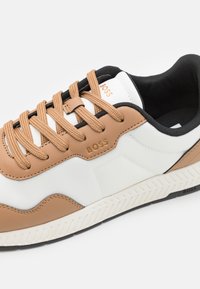Bőr és szövet sneaker fehér, barna és fekete részletekkel. Jellemzője a texturált felsőrész, a kerek fűzők és a mintázott gumi talp. A márkajelzés látható.