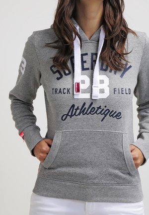 Kobieta w szarej bluzie Superdry z białymi sznurkami, przednią kieszenią i napisem "Superdry 23 Track Field Athletique" w granatowym i białym kolorze.